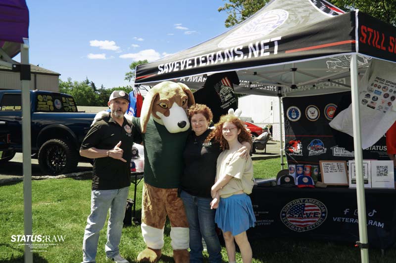 People posing with mascot in front of veterans tent display